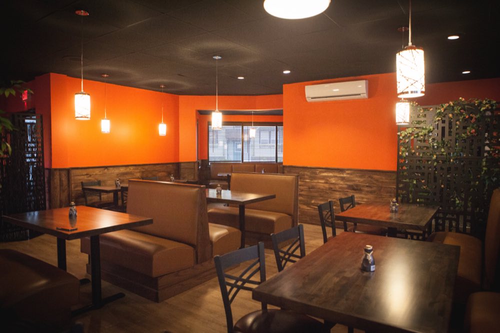 The front dining room seen from the entrance, with booths and tables against orange walls over wood-plank wainscoting and a window onto the street.