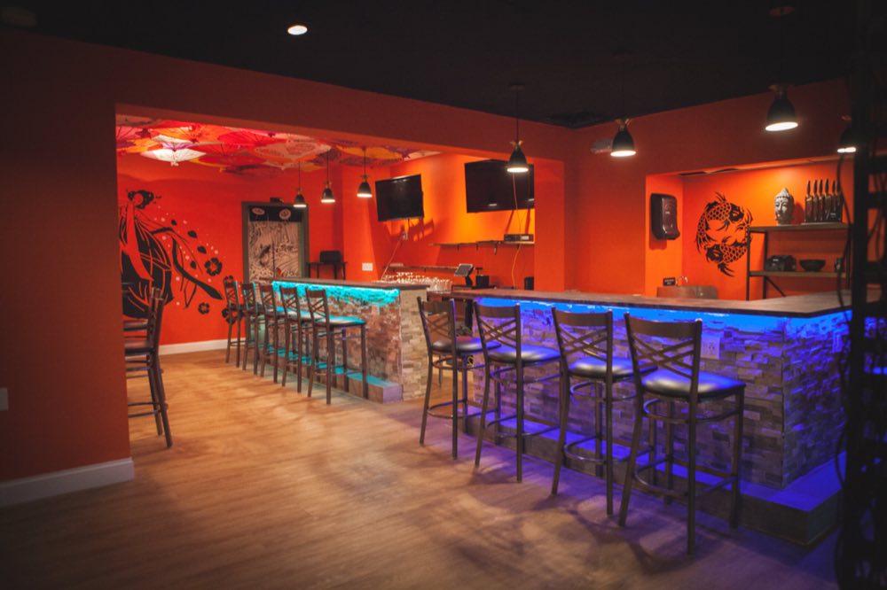 The bar room: a long stone-faced bar lit blue from beneath with high-backed stools, two wall-mounted TVs, and the parasol-covered alcove beyond.