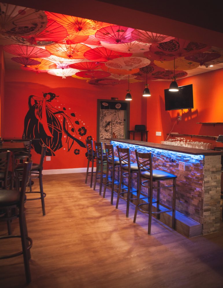 A ceiling completely covered in open paper parasols in red, pink and cream above the bar, with a large black brush-drawn geisha mural on the orange wall and wine glasses set out along the blue-lit stone bar.